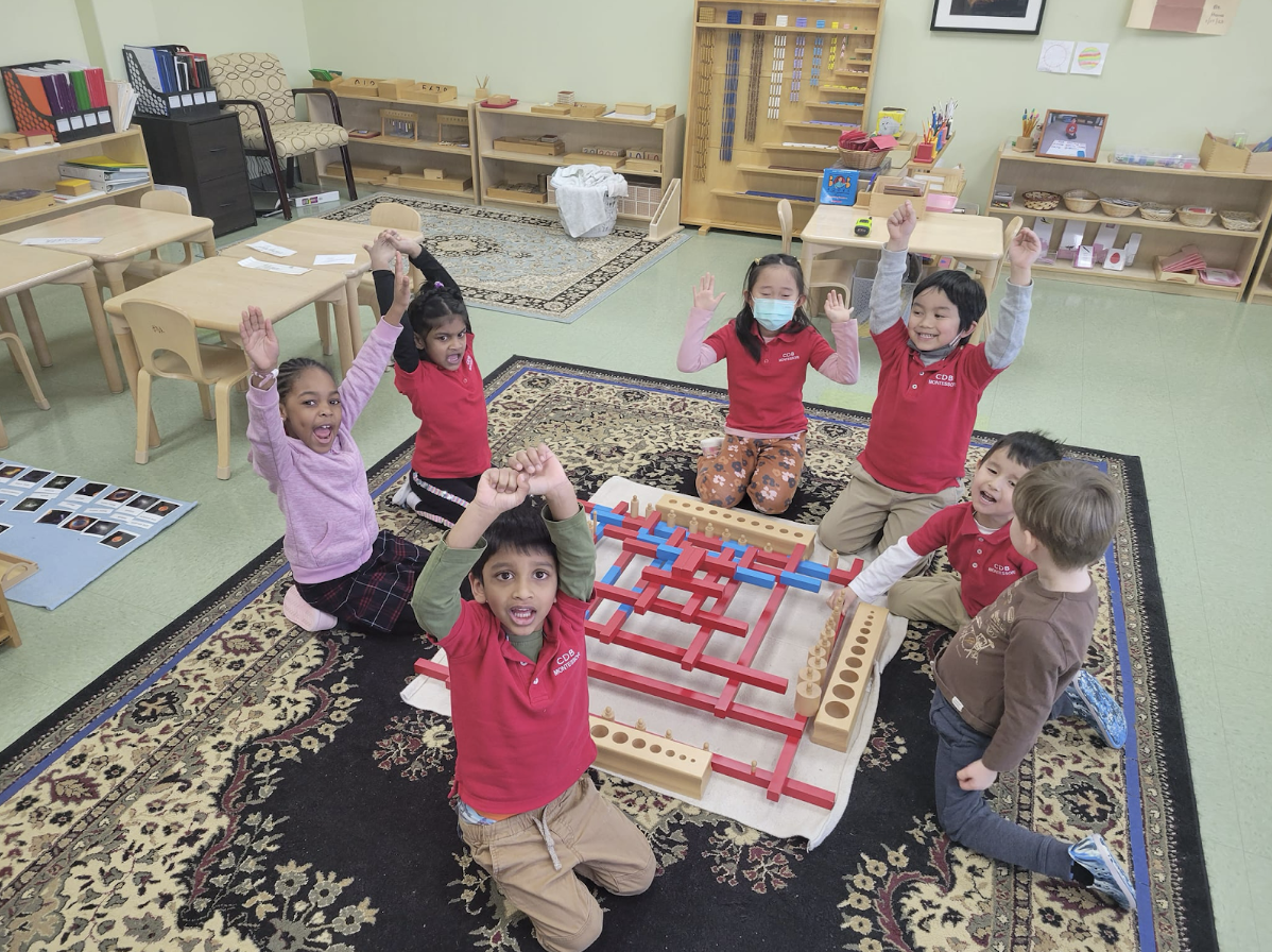 A group of children in a classroom raising their hands while sitting on a rug with educational toys.